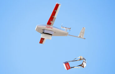 A Zipline fixed-wing drone in the air drops a package