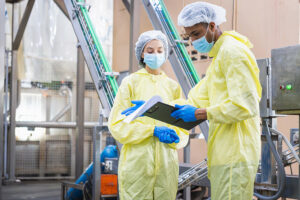 Diverse factory workers in yellow PPE suits checking production data with clipboard in modern industrial facility, Quality control inspector wearing masks and hairnets working in food or pharma plant