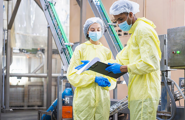 Diverse factory workers in yellow PPE suits checking production data with clipboard in modern industrial facility, Quality control inspector wearing masks and hairnets working in food or pharma plant