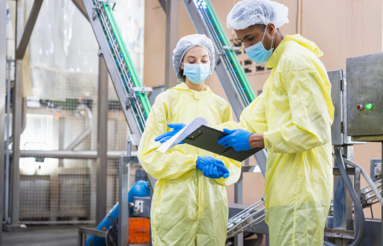 Diverse factory workers in yellow PPE suits checking production data with clipboard in modern industrial facility, Quality control inspector wearing masks and hairnets working in food or pharma plant
